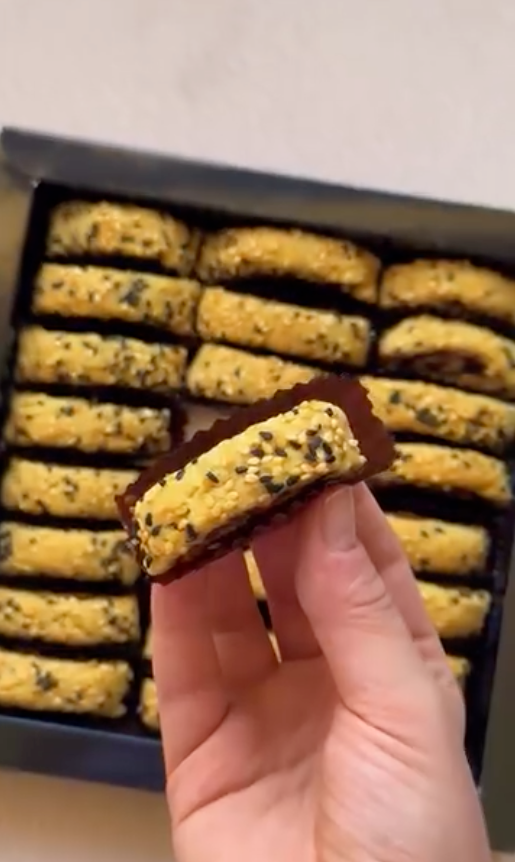 A hand holding a single square date cookie with a dark chocolate base and golden-yellow crumb top speckled with black seeds, with more cookies visible in a black tray in the background.