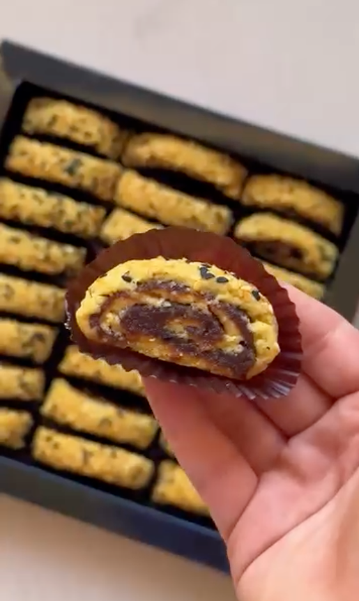 A hand holds a halved yellow pastry with a date swirl, in front of a box of similar pastries.