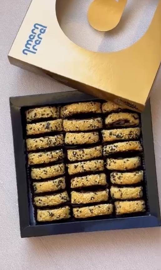 An open black square box containing multiple rows of golden-brown chocolate chip cookies, with a gold and cream branded box lid placed behind it on a light surface.