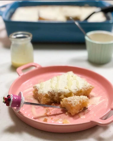 A close-up of a pink plate holding a slice of cake or pastry topped with white frosting, with a fork resting on the plate. A blue baking dish, a small glass jar, and a cup are blurred in the background on a light surface.