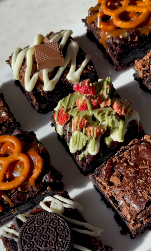 An assortment of chocolate brownies topped with white chocolate drizzle, pretzels, strawberry cream, green chocolate drizzle, and Oreo cookies, arranged on a white plate.