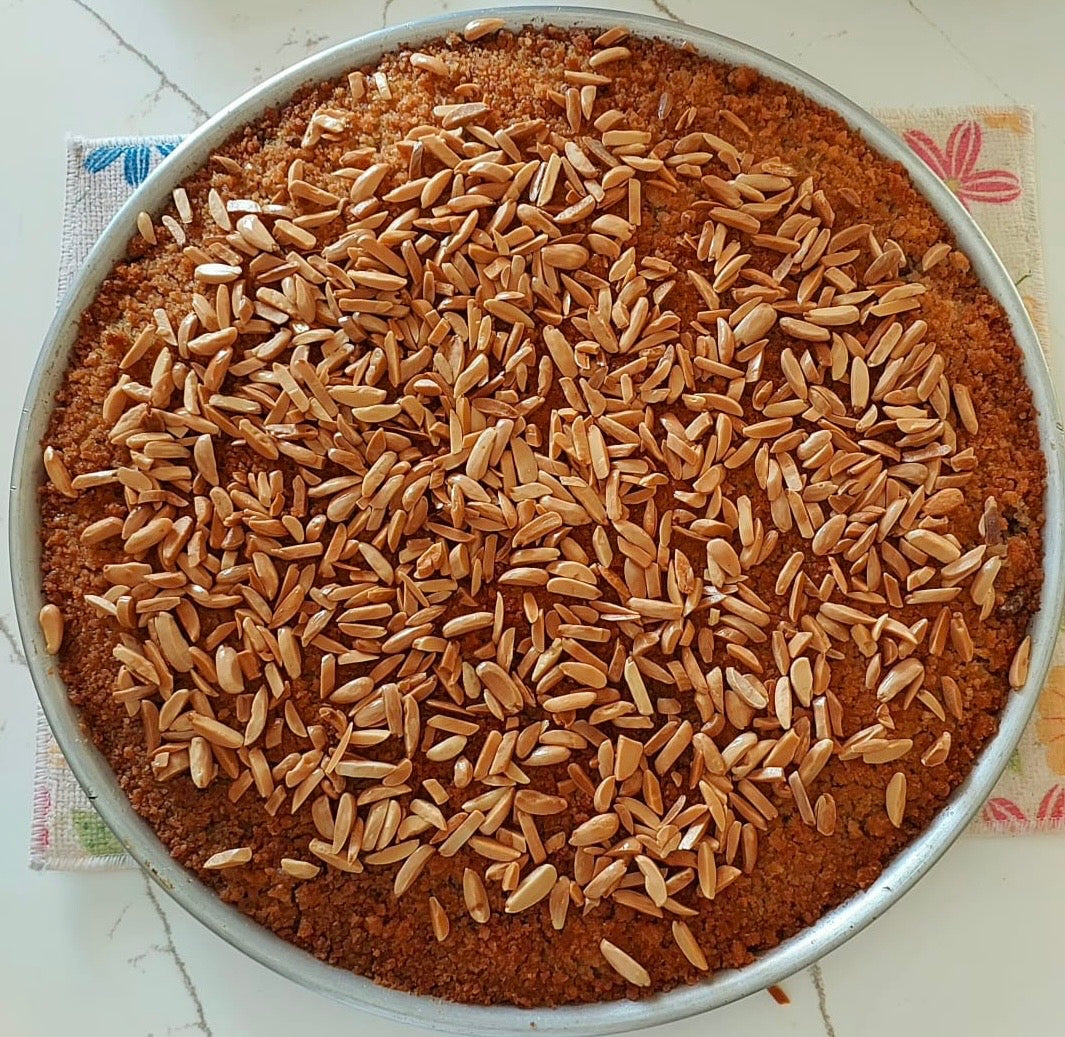 A round metal baking pan filled with a brown crumb base and topped with a dense layer of whole wheat or oat-like grains, photographed from above on a light surface with a patterned cloth underneath.