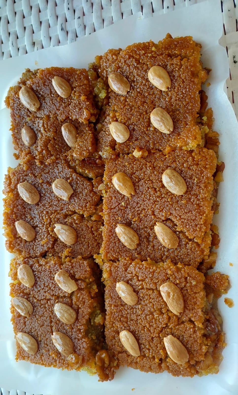 Six rectangular pieces of golden-brown sweet pastry or cake, each topped with a few whole almonds, arranged on a white plate over a woven light surface.