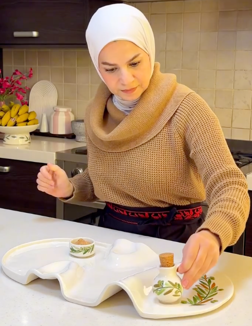 A woman wearing a beige cowl-neck sweater and a white hijab is shown in a kitchen setting, placing a small white ceramic bottle with a cork stopper and green botanical illustrations onto a white wavy serving tray.