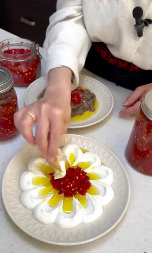 A close-up of a person preparing a dish, placing a piece of ginger into a white cream flower-shaped arrangement on a plate with red chili paste and yellow oil. Several glass jars filled with red chili paste are visible on a white countertop.