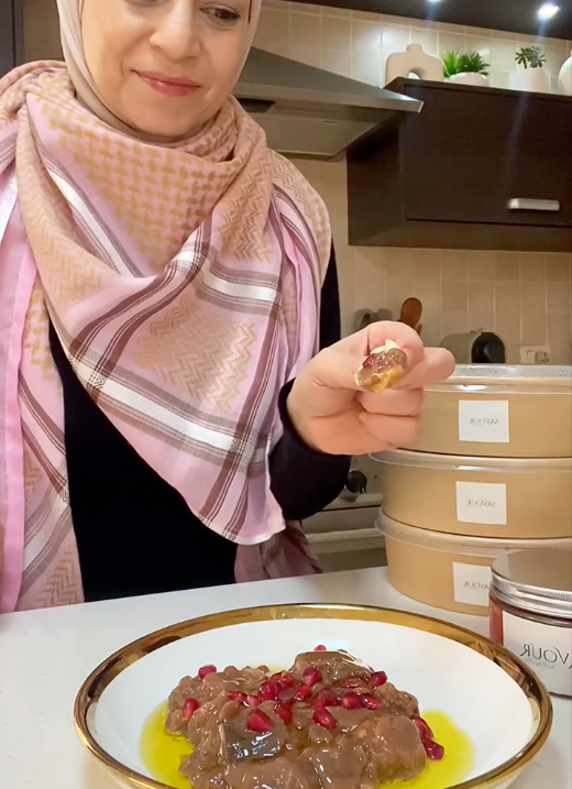 A woman wearing a pink and beige patterned hijab holds a small piece of food in her hand, with a plate of cooked meat in oil and pomegranate seeds in the foreground and stacked containers in a kitchen setting behind her.
