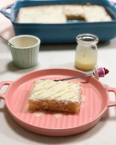 A square slice of golden-brown cake or bread dusted with powdered sugar and drizzled with syrup, served on a pink plate with a small spoon. A blue baking dish, a white ramekin, and a small glass bottle of yellow sauce are arranged on a white surface in the background.