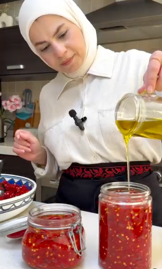 A woman in a white shirt and black skirt with a red patterned waistband is pouring golden olive oil into two glass jars filled with chopped red chili peppers. The setting is a kitchen with dark cabinets and a countertop.