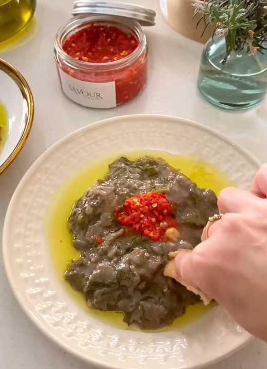 A close-up of a hand dipping a piece of bread into a plate of Sumaqiyah in oil, topped with a red chili paste, with a jar of red sauce in the background.